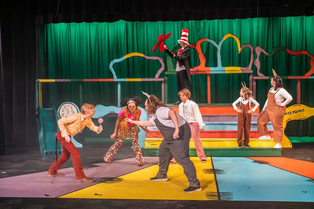 Performers in colorful costumes act in a theatrical stage production with vibrant green and orange lighting elements.