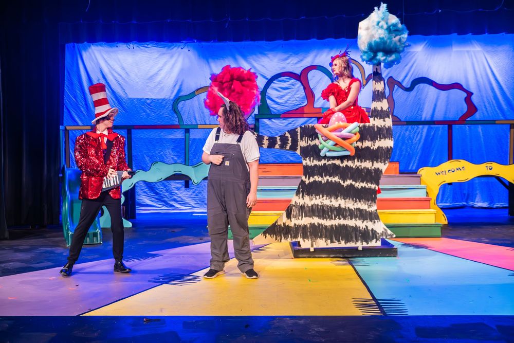 A theatrical production scene with vibrant red and blue lighting features performers in colorful costumes on stage.