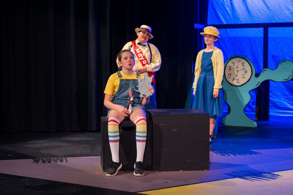 Performers in colorful costumes act on stage during a theatrical production with blue lighting and set pieces.