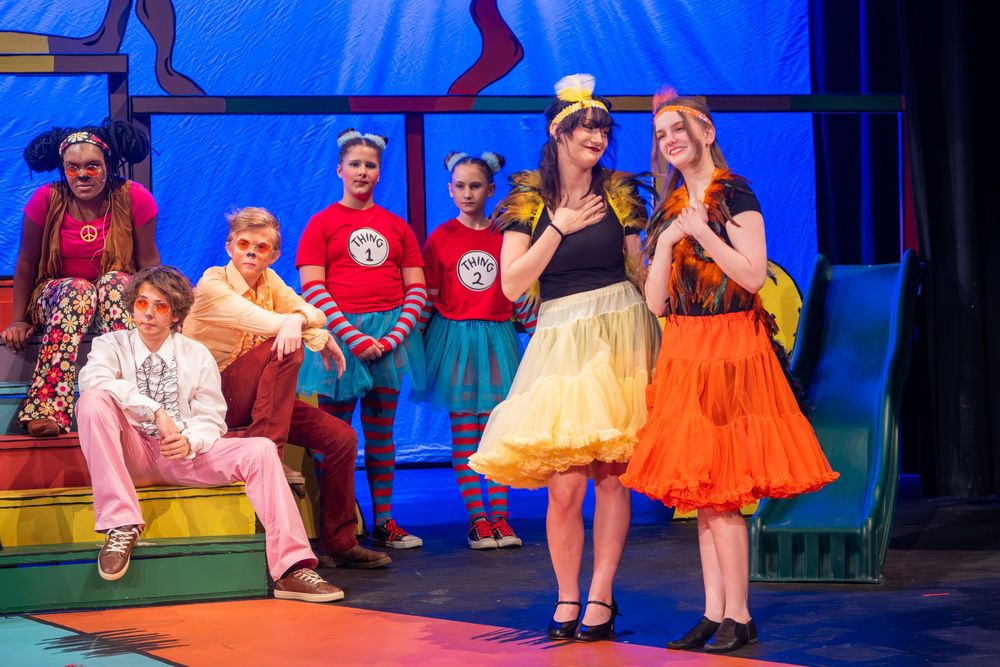 Colorful stage performance featuring actors in retro-style dresses and casual wear against a bright blue backdrop.