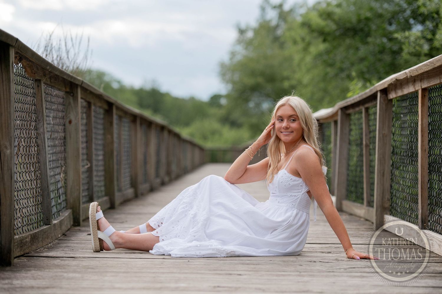 A person in a white flowing dress poses gracefully on a wooden bridge walkway surrounded by greenery.