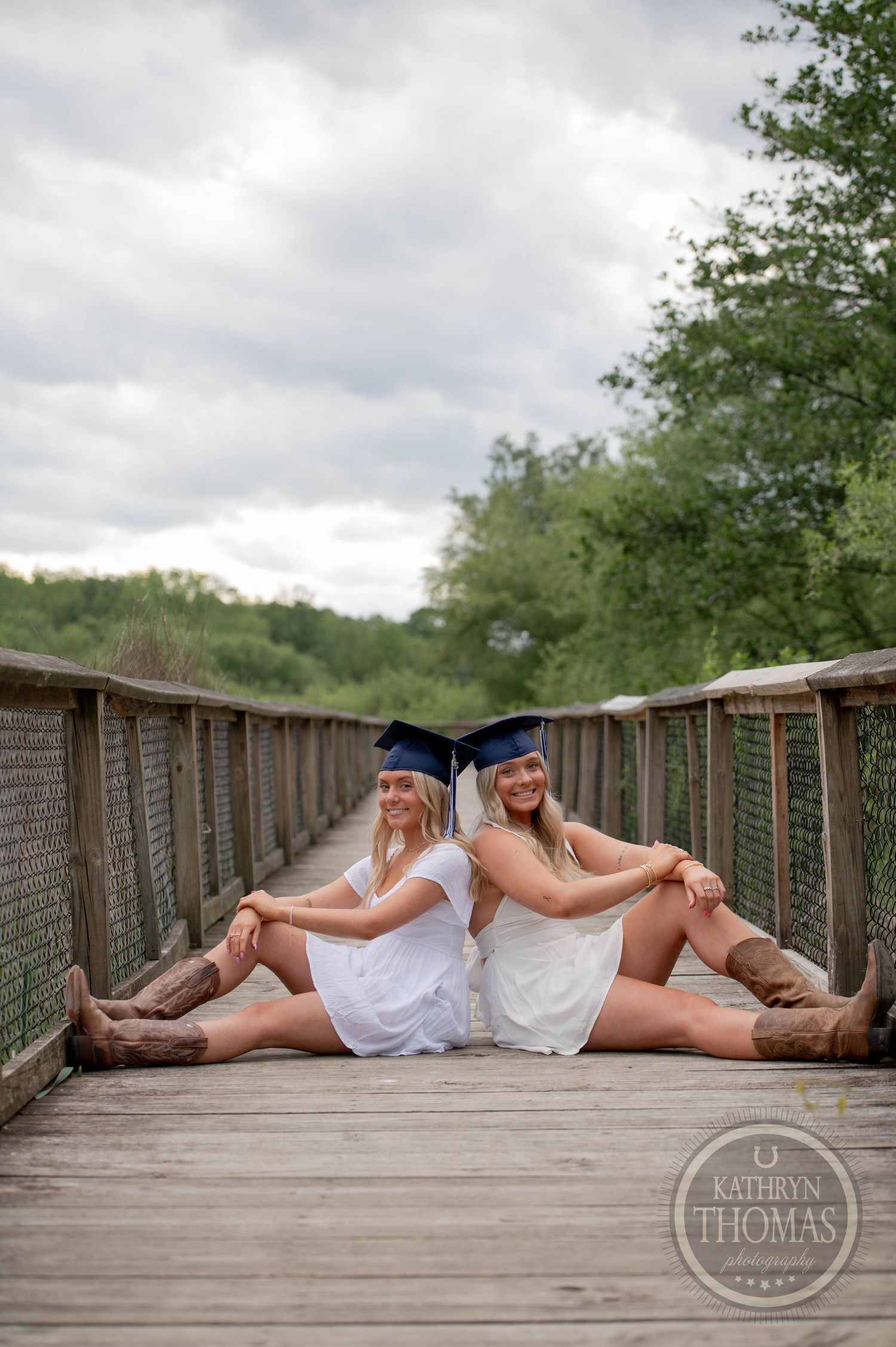 Two graduates in white dresses and graduation caps sit back-to-back on a wooden bridge path through nature on a cloudy day.