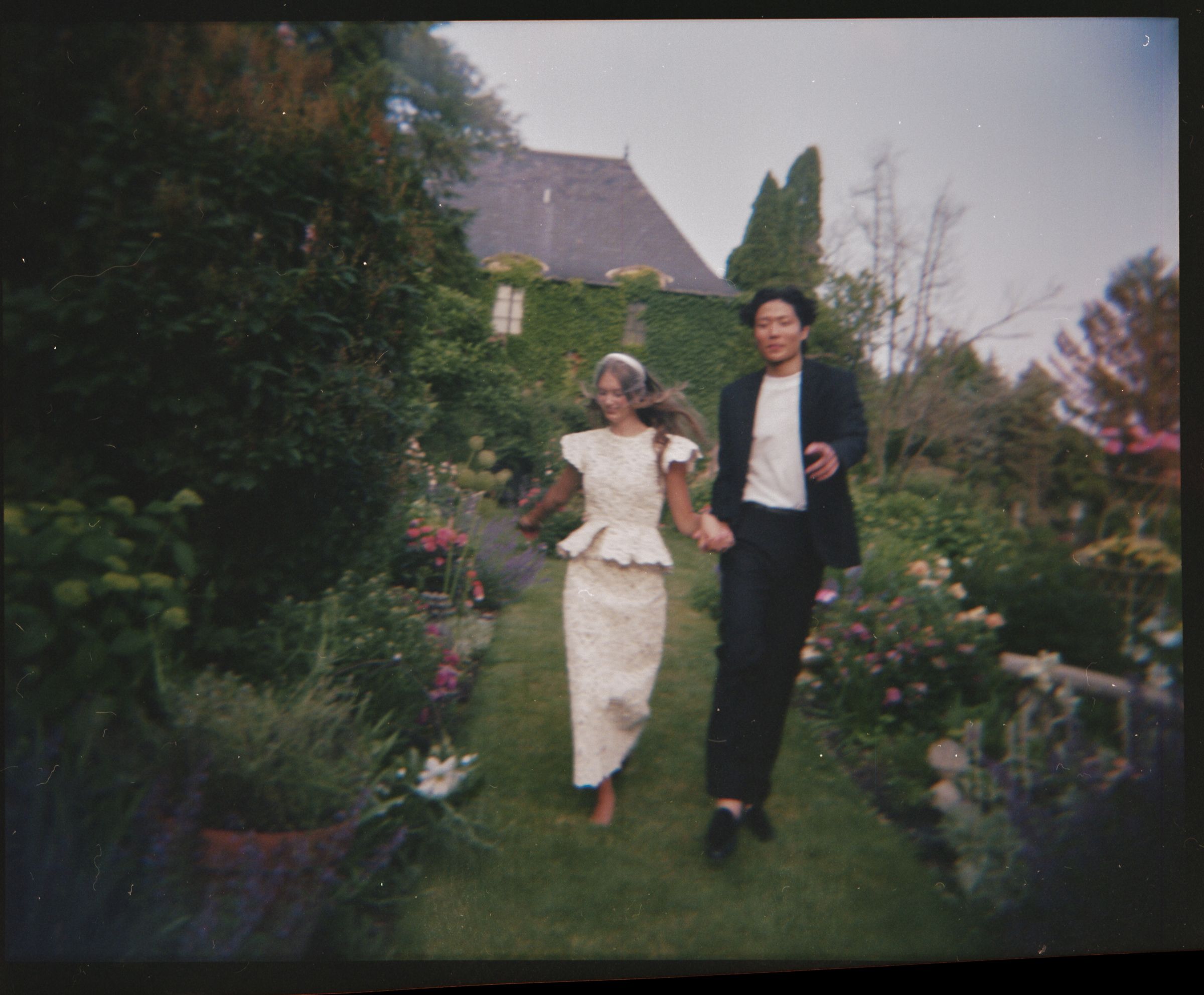 A couple walks together through a lush garden path with flowers, dressed formally in vintage style attire.