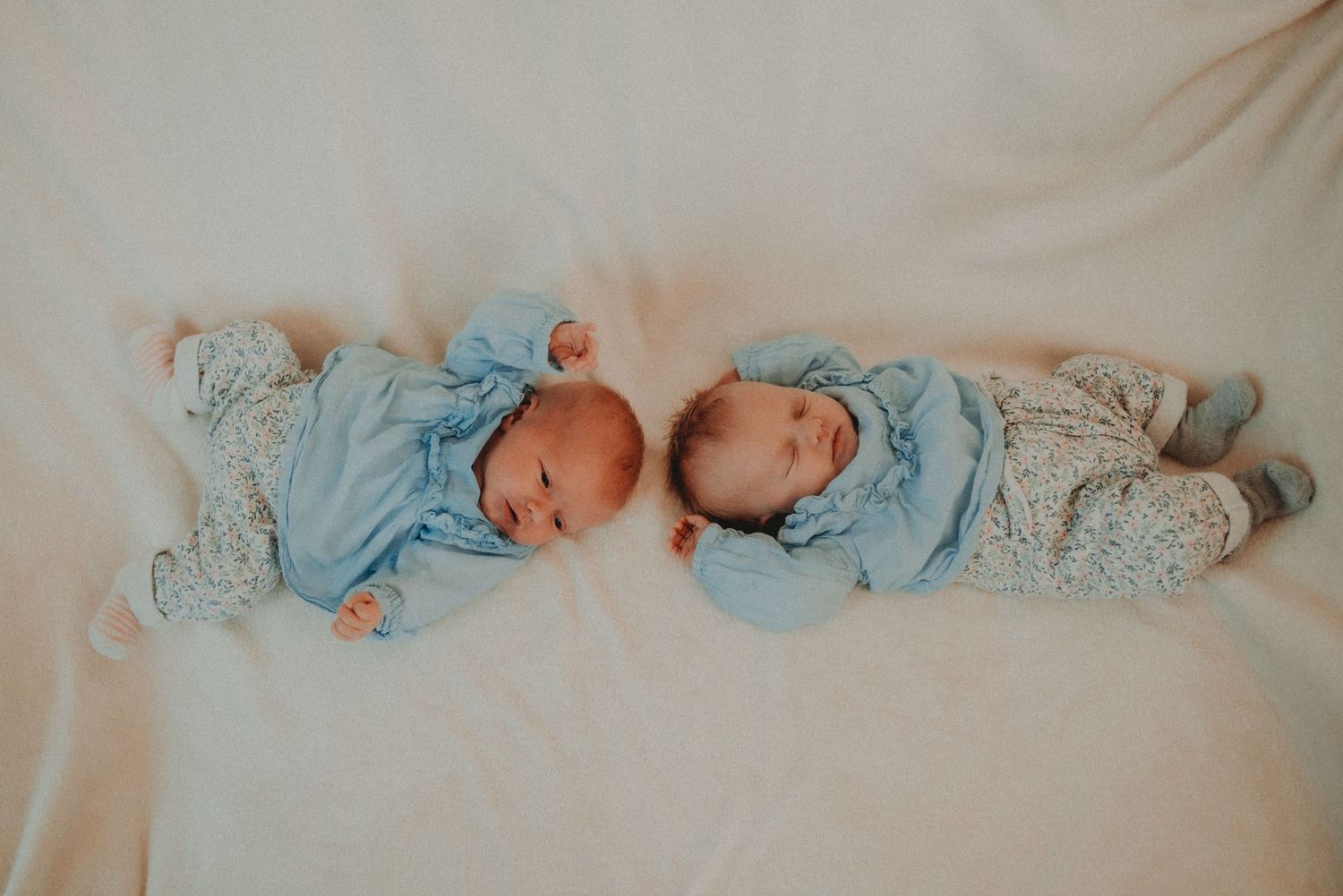 Two sleeping newborn babies wearing matching light blue outfits lying side by side on a white blanket.