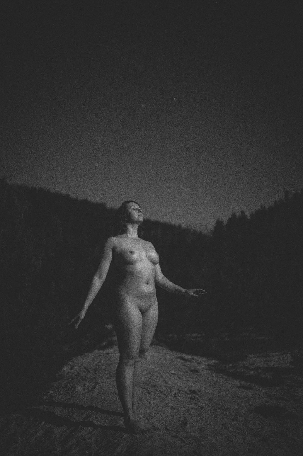 Artistic black and white nude photograph of person standing on rocky ground looking upward at night sky with trees in background.