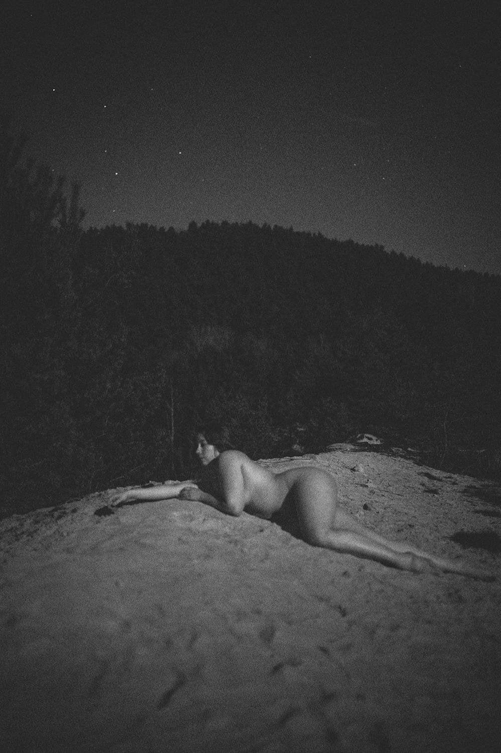 Black and white photo of a nude figure reclining on a rock formation at night with hills and stars in the background.