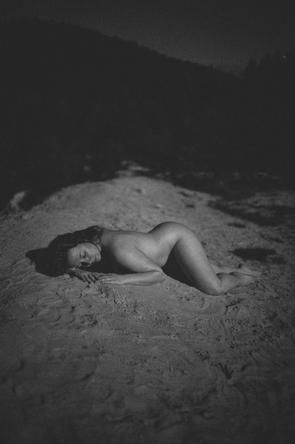 A black and white artistic nude photograph of a person reclining on sand in nature.