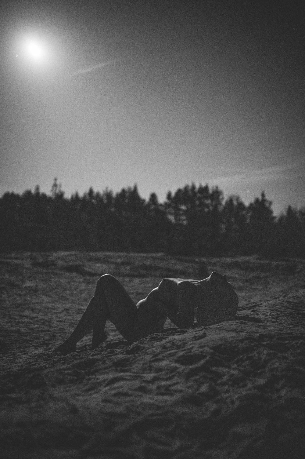 A silhouetted figure lies on sandy ground beneath a bright moon and starlit sky in black and white.