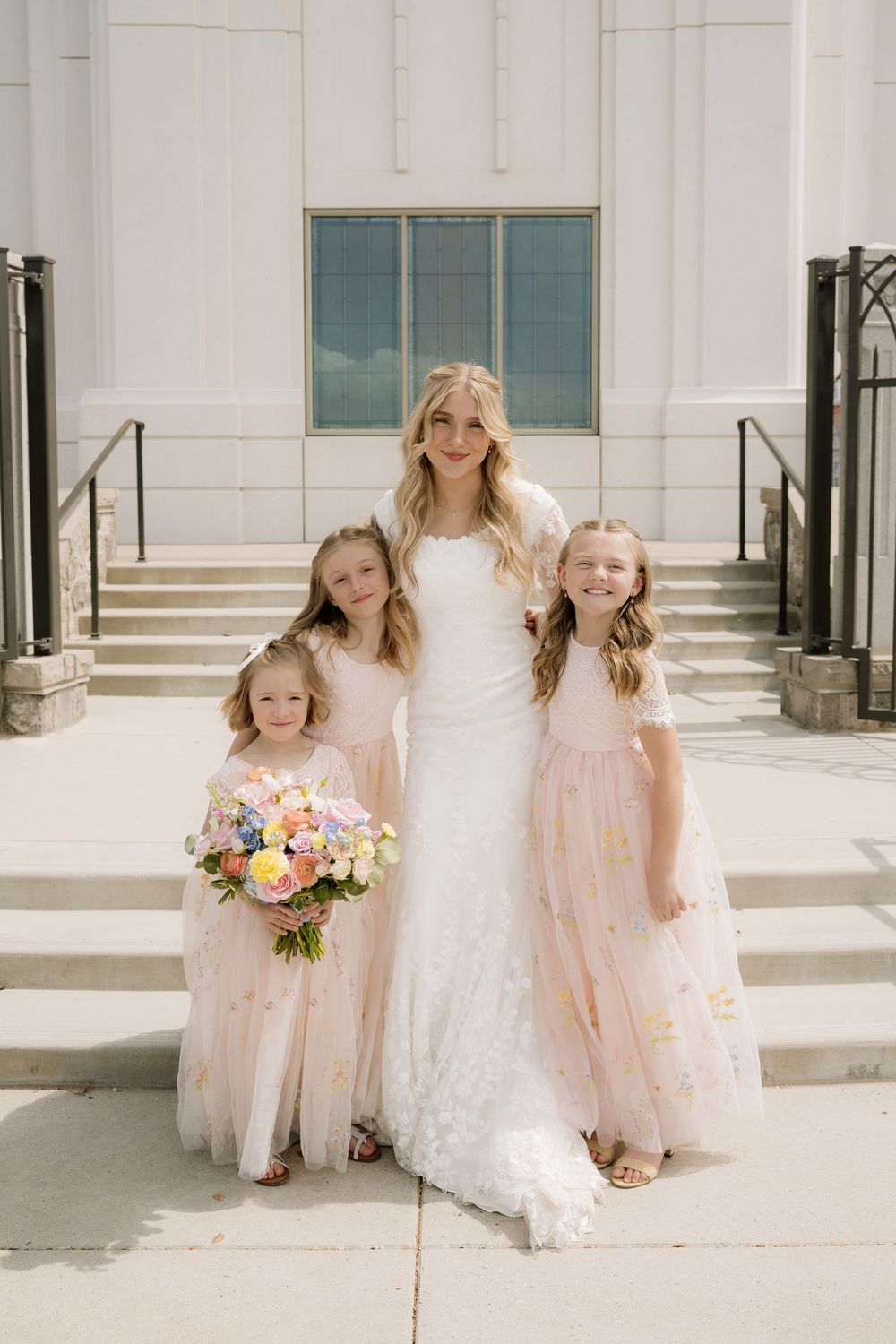Bride in white lace gown poses with three flower girls wearing blush pink dresses.