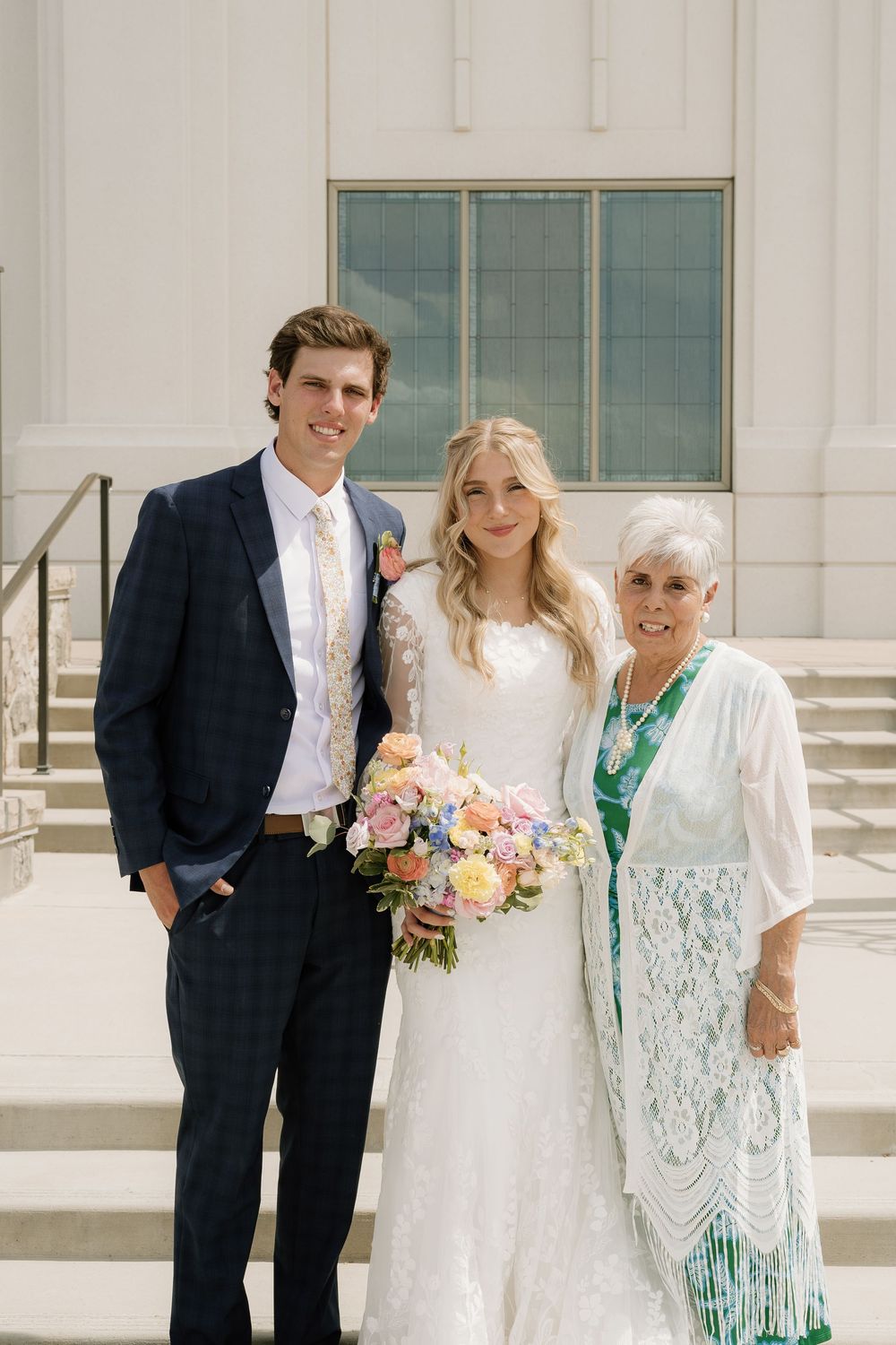 Wedding couple in formal attire pose with elder family member on church steps.