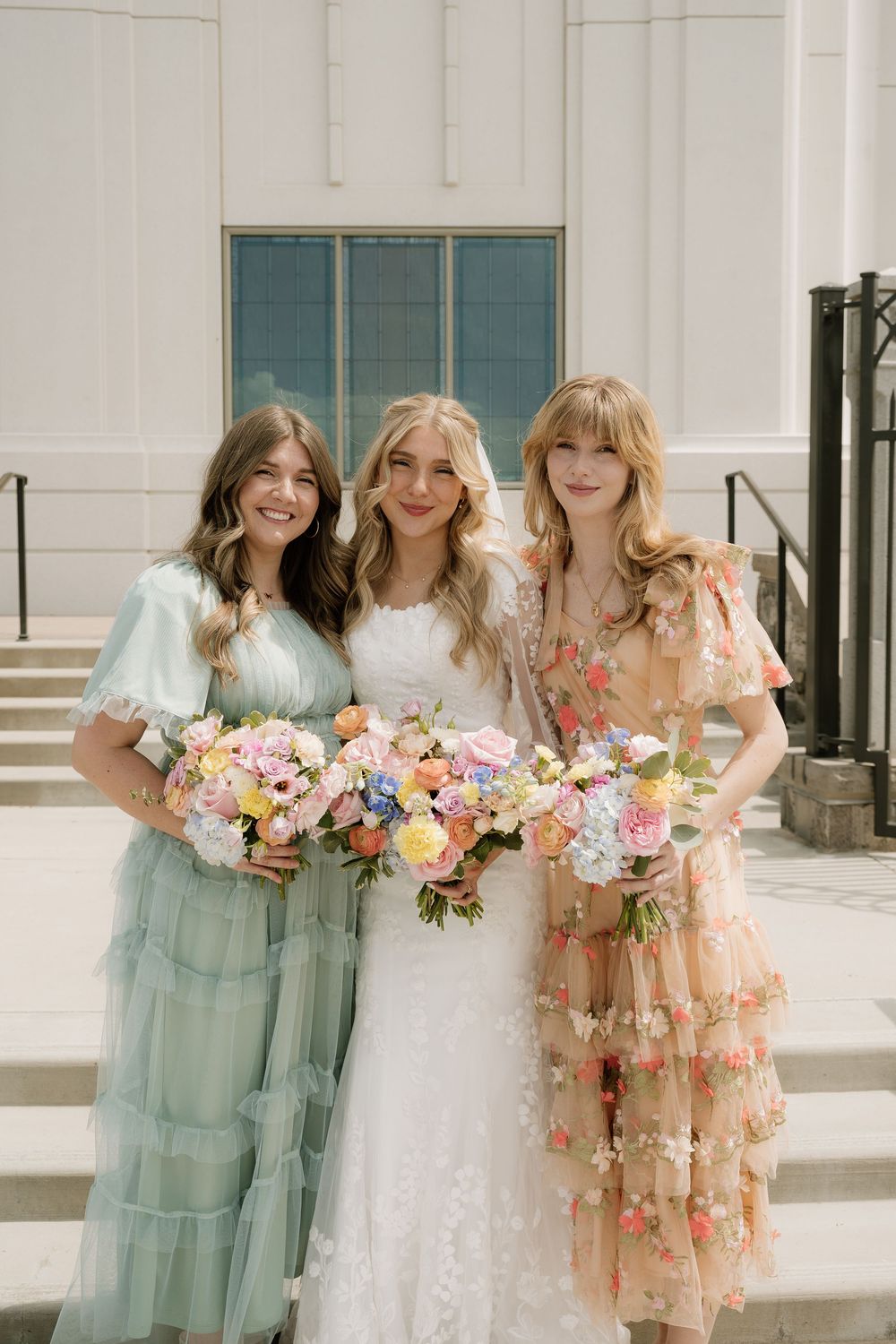 Three bridesmaids in pastel dresses hold spring flower bouquets on church steps.