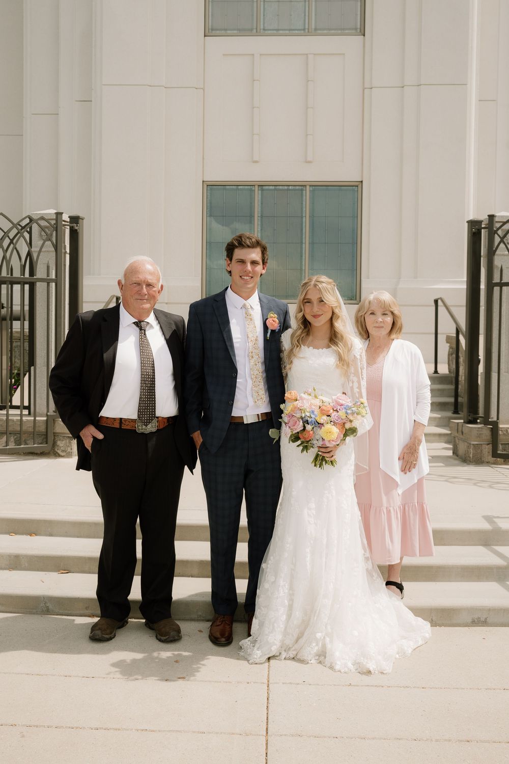Four people stand together on white stone steps outside a church building at a wedding ceremony.