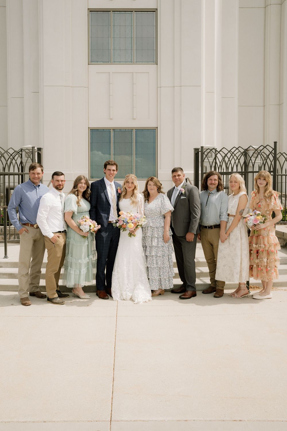 Wedding party in pastel and neutral clothing poses on white stone steps outside a formal building.