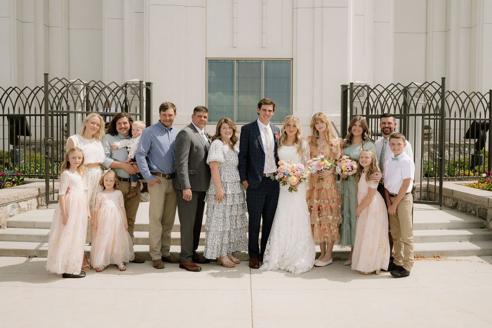 A larger wedding group stands together on white steps with decorative black iron fencing behind them.
