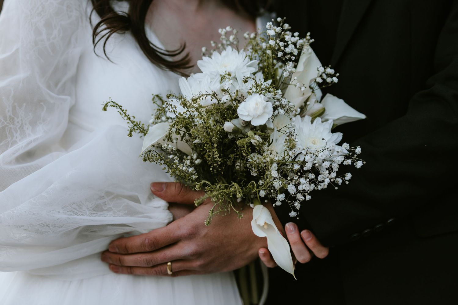 Close-up view of a wedding bouquet with white flowers and delicate greenery held between hands with a wedding band.