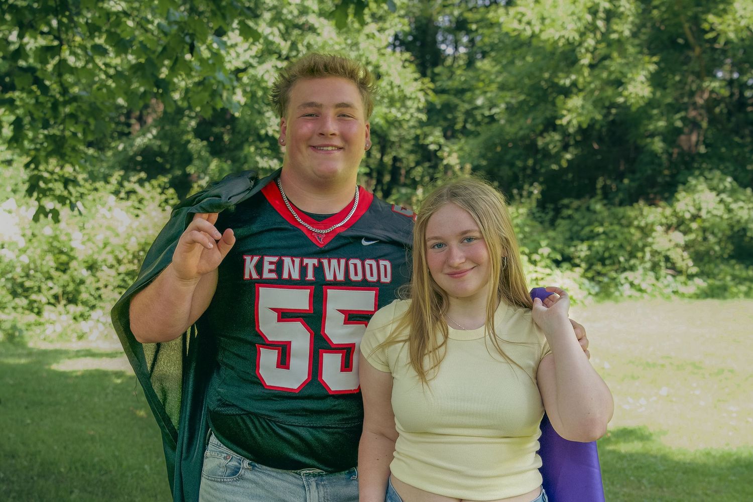 Two young people in a sunny outdoor setting wearing casual attire and making peace signs while smiling at the camera.