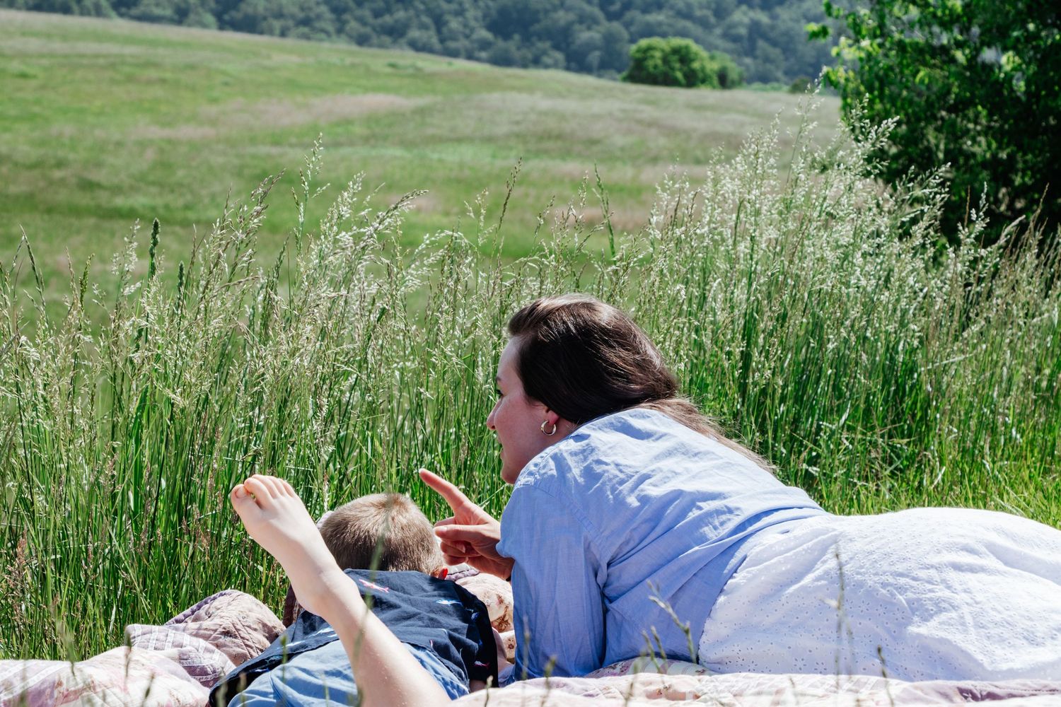 Mother & son laying on a quilt in a grassy field, pointing at something together while enjoying nature.