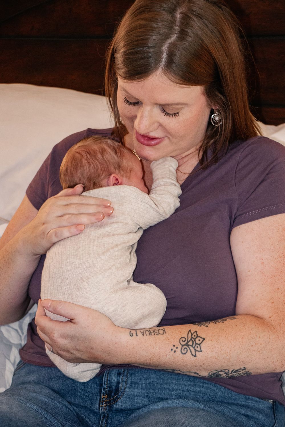 Mother sitting on a bed, lovingly holding her sleeping newborn against her chest while smiling down at them.
