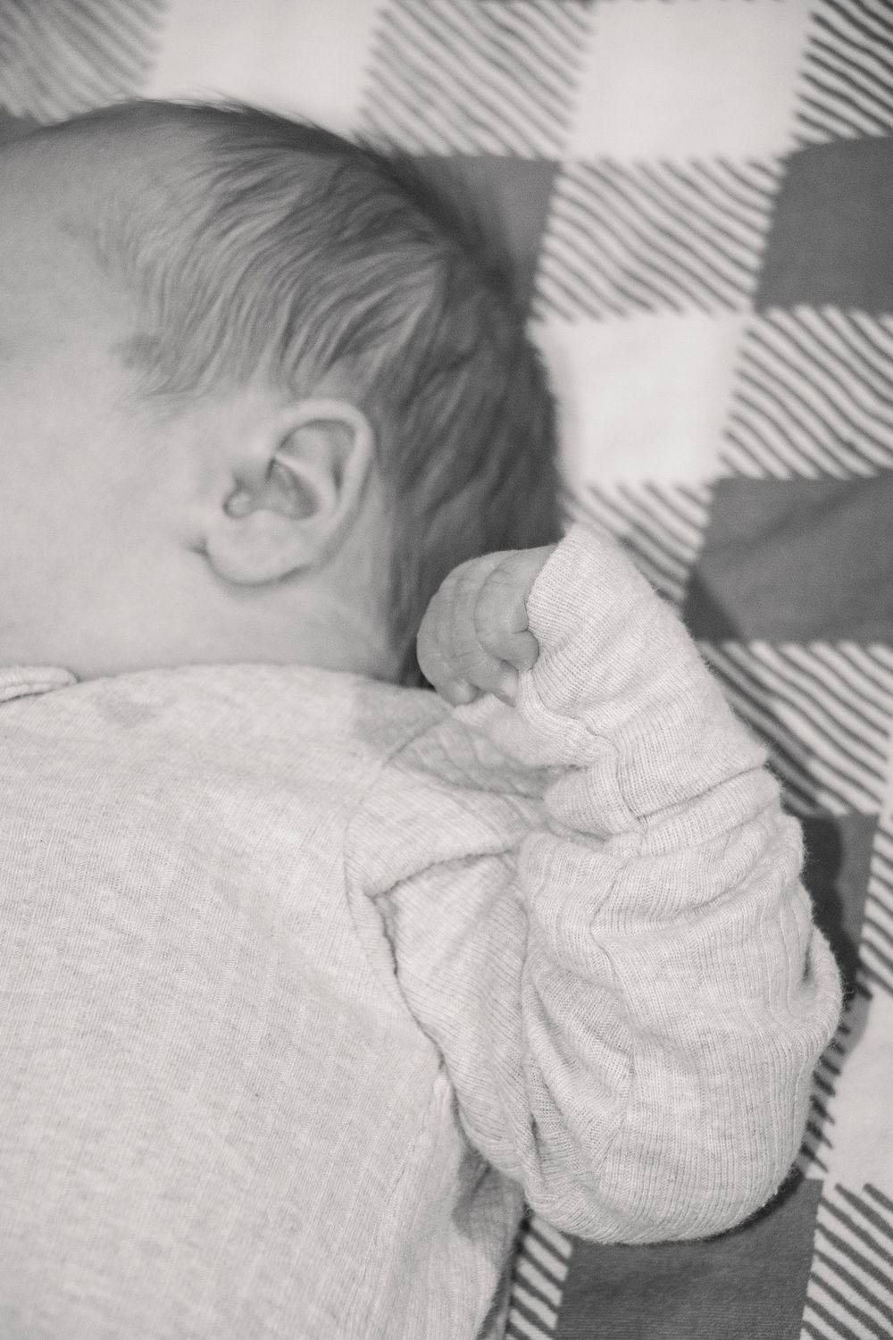 Black & white photo of a newborn resting peacefully on a checkered blanket, showing only part of the face and hand.