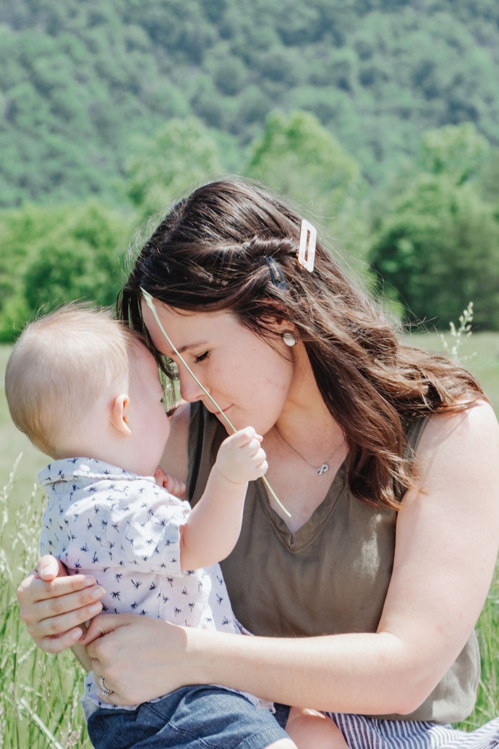 Tender moment of a mother and baby sitting in a field, touching foreheads while the baby holds a piece of grass.