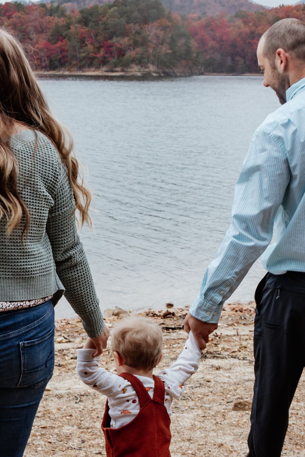 Mom, Dad, and toddler holding hands while facing a calm lake surrounded by fall trees, seen from behind.