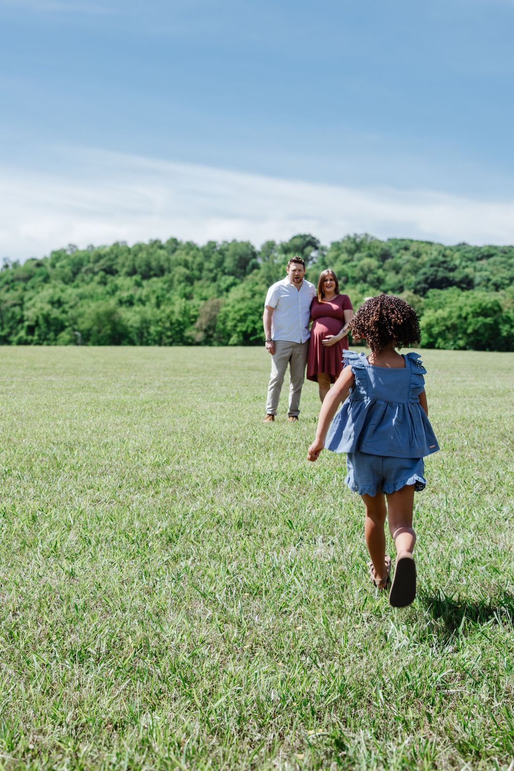 Young girl runs across a sunny field toward her smiling parents, with the mother visibly pregnant and greenery in the background.