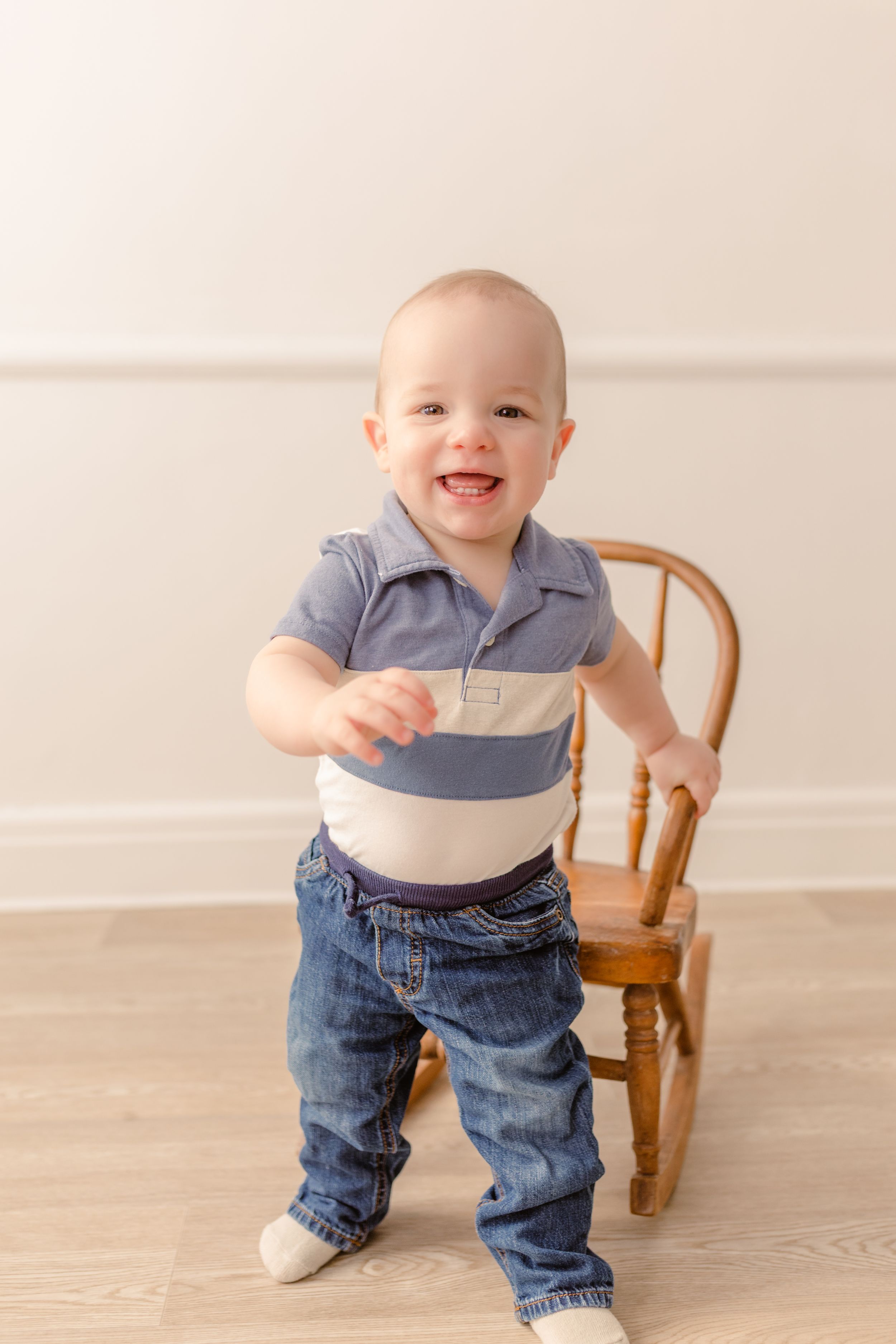 Smiling baby in blue shirt and jeans standing near wooden chair.