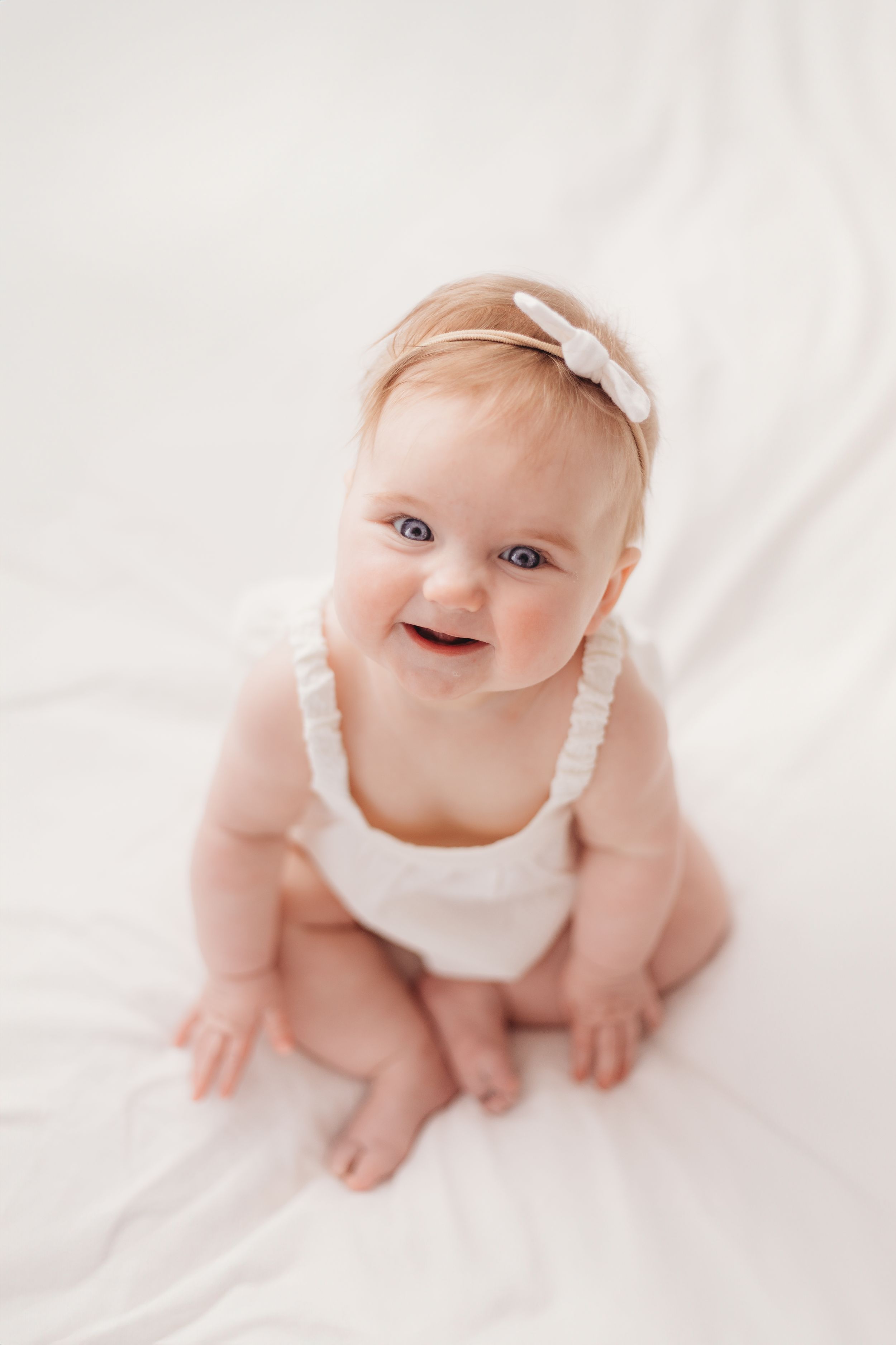 Smiling baby wearing white bow headband and tank top on white background.
