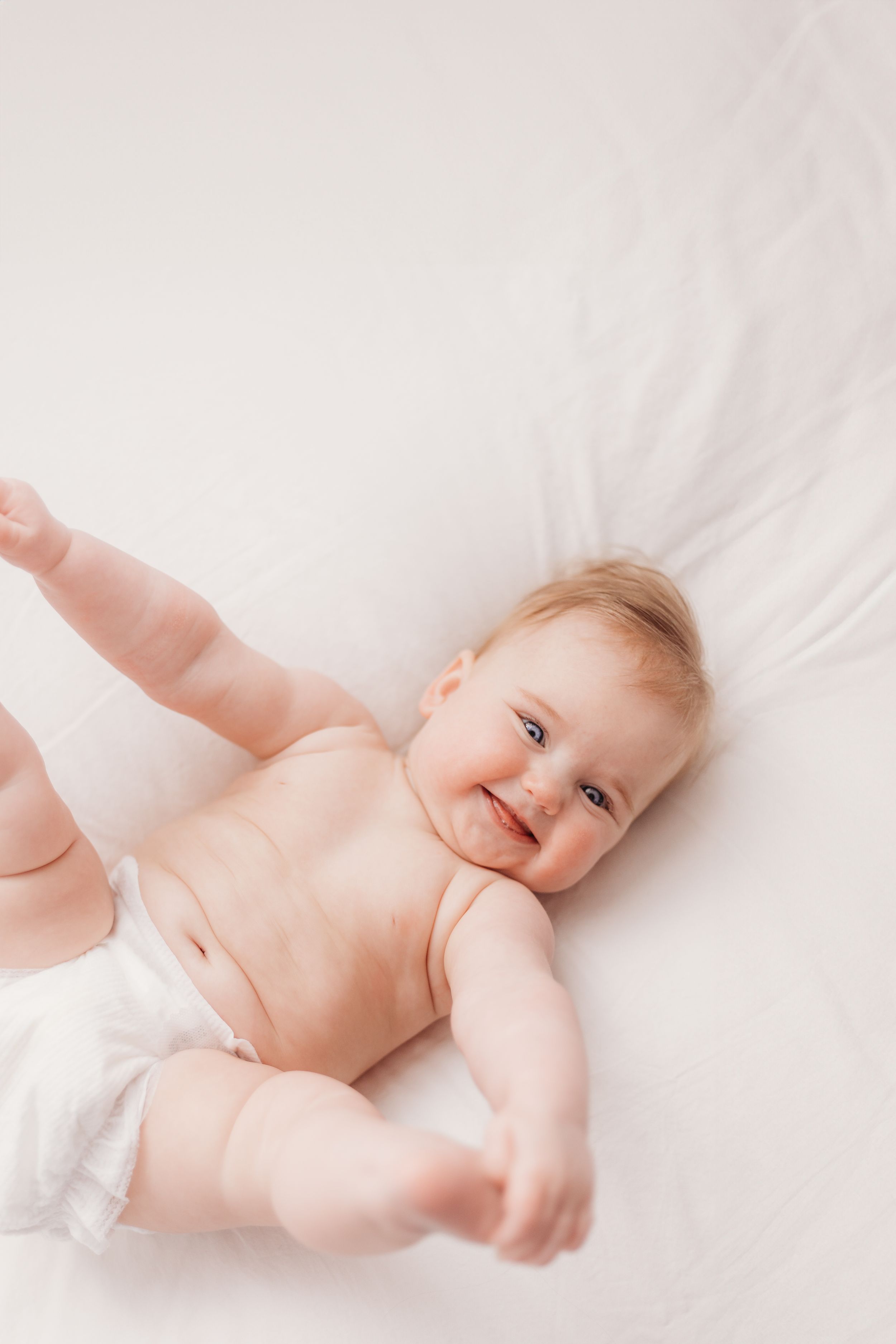 Joyful baby lying on white bedding with arms and legs stretched out.