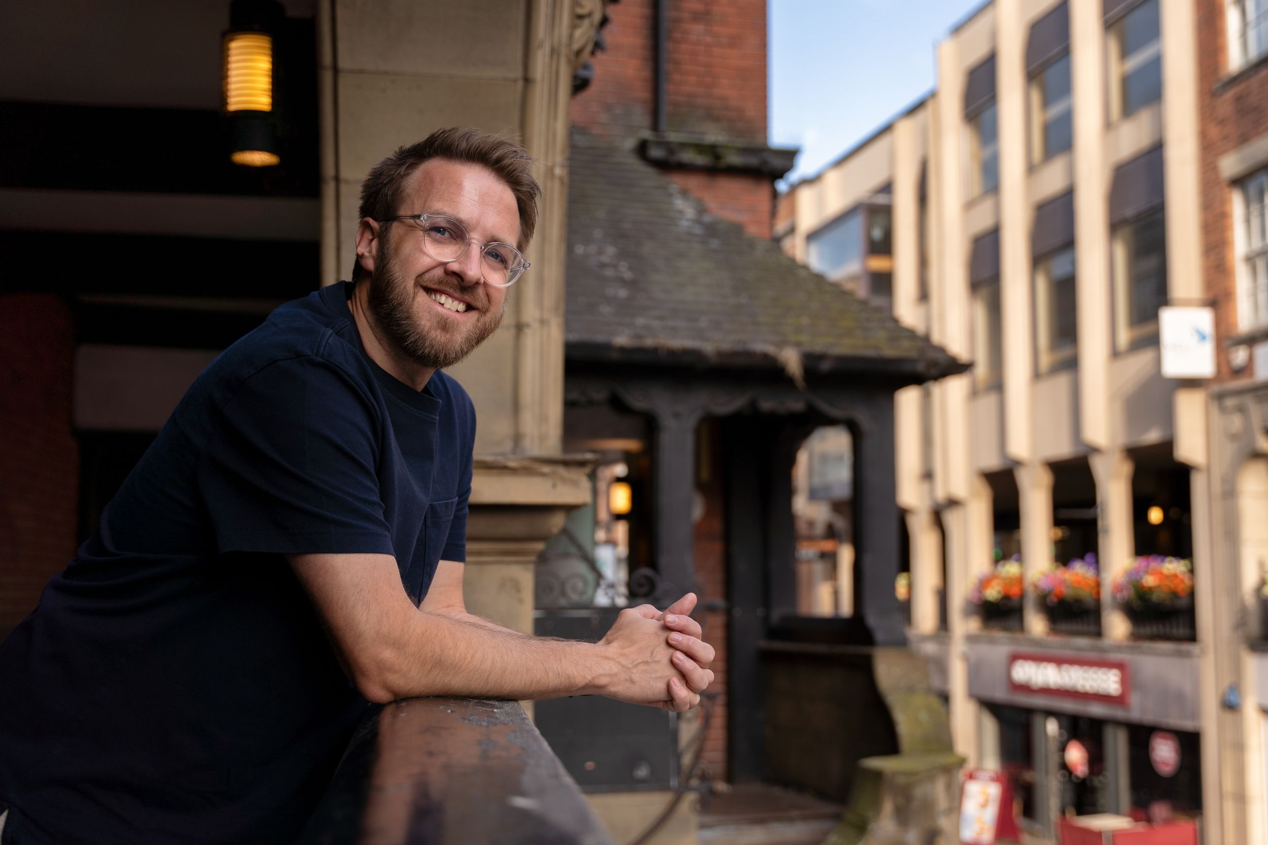 Life Coach wearing a navy shirt smiles while leaning on a railing with brick buildings and an amber light visible in background.