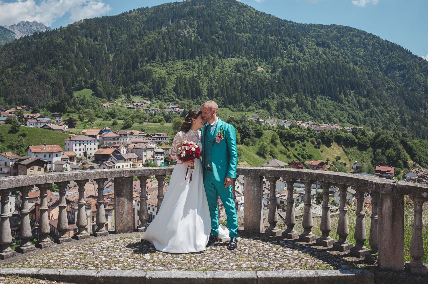 Couple in wedding attire embraces on stone balcony overlooking mountain village in Italian Dolomites on sunny day.