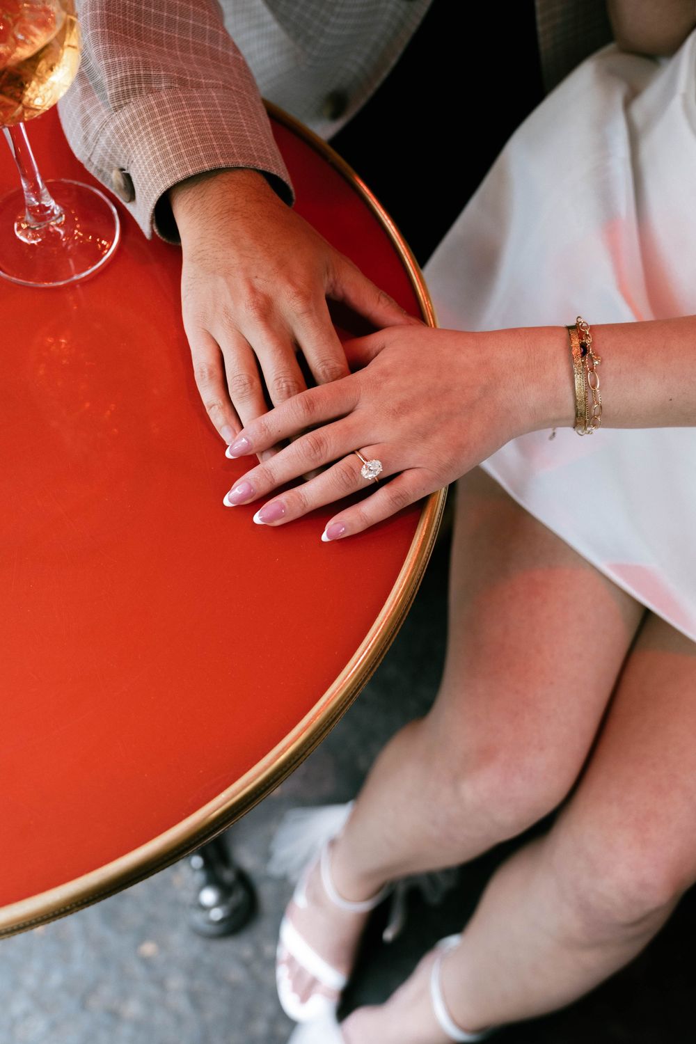 Hands with engagement ring and bracelet resting on a round red table while wearing white sandals.