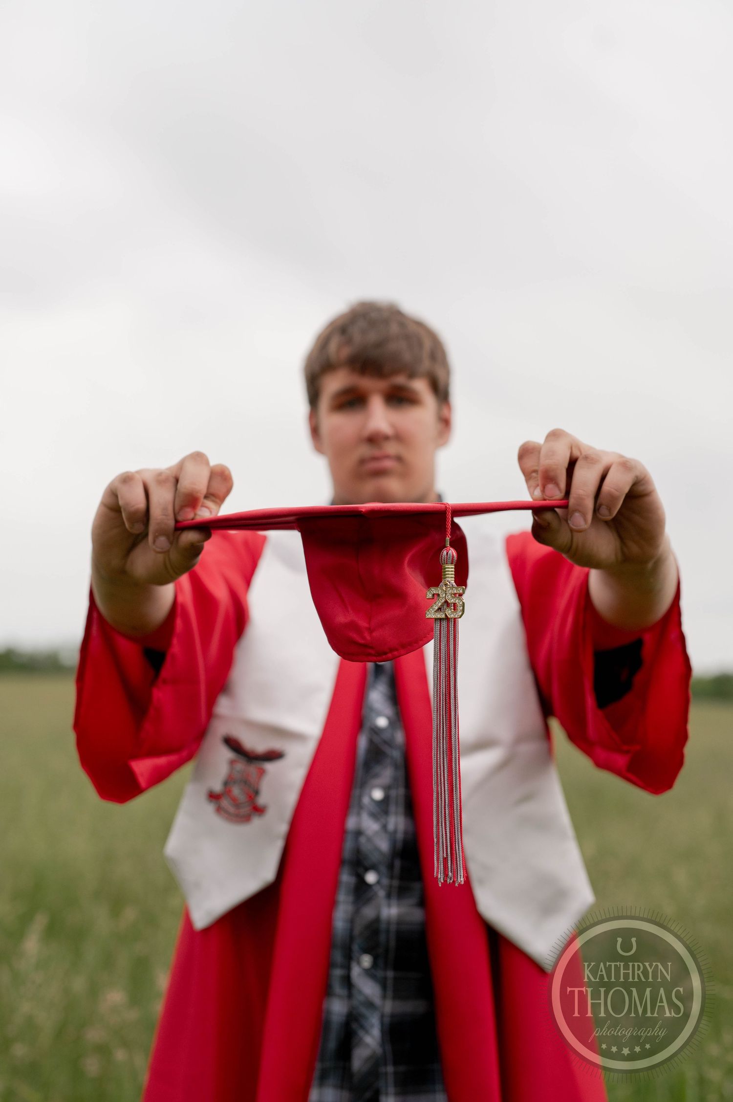 Graduate student in red and white academic regalia holding up tassel against blurred outdoor background.