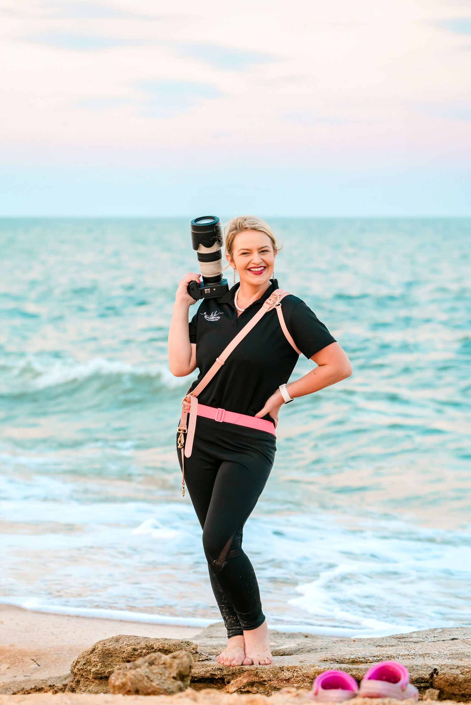 Photographer with camera stands barefoot on rocky beach with turquoise ocean waves in background.