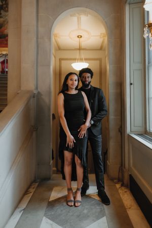 A couple dressed in formal black attire poses elegantly in an arched hallway with classic architectural details.