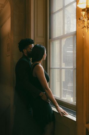 A couple stands together in warm light by a vintage window with ornate trim and chandelier glow in the background.