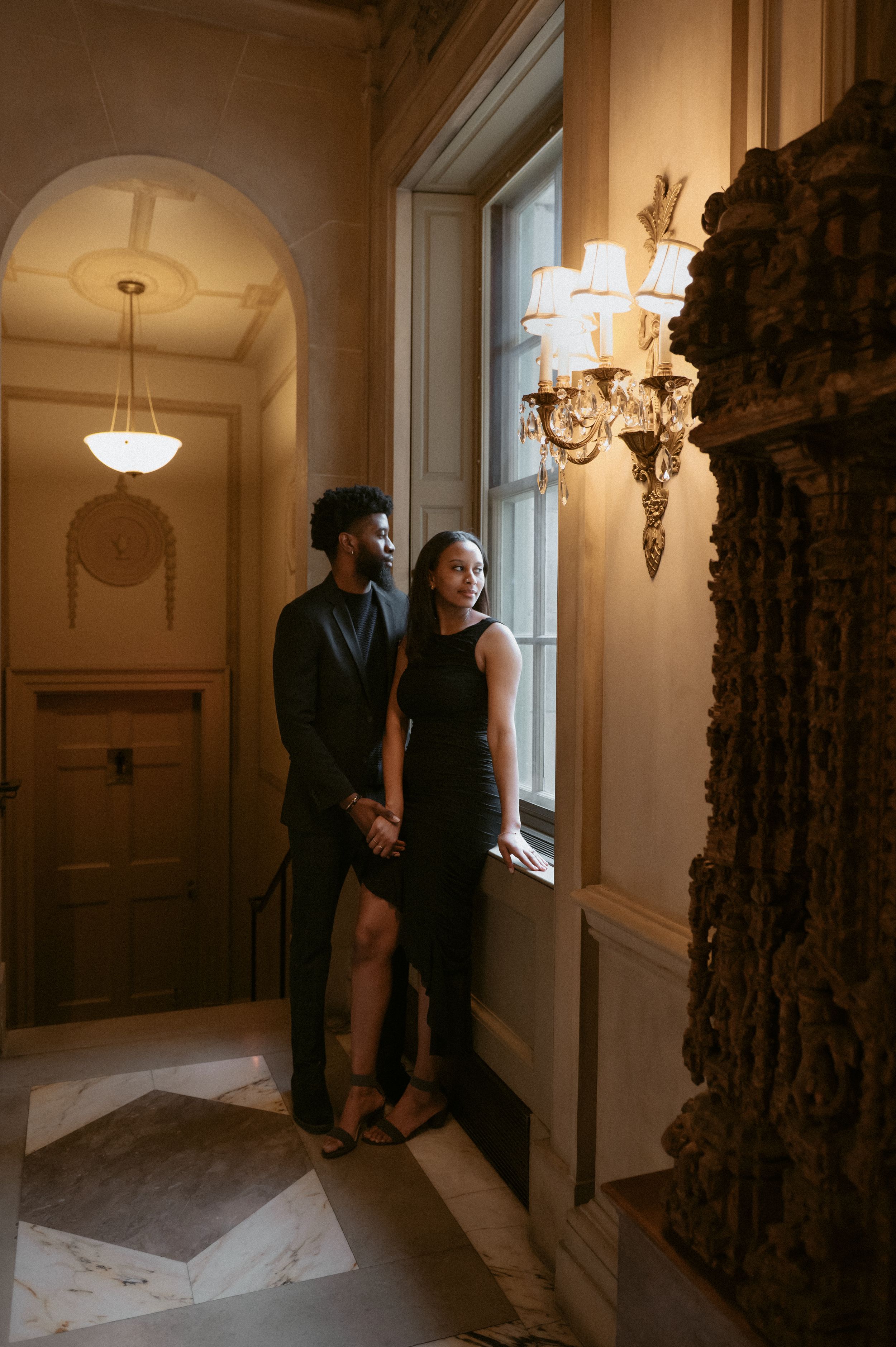 A couple shares an intimate moment standing by an ornate window in an elegant hallway with classical architecture.