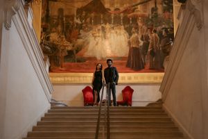 Two wedding photographers stand on elegant white stairs with red chairs and a large historical painting in the background.