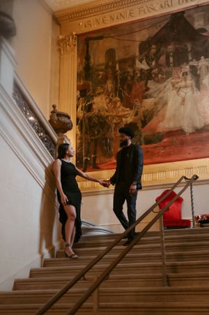 Two people in elegant black outfits stand on a grand staircase with ornate artwork and golden framing on the wall.