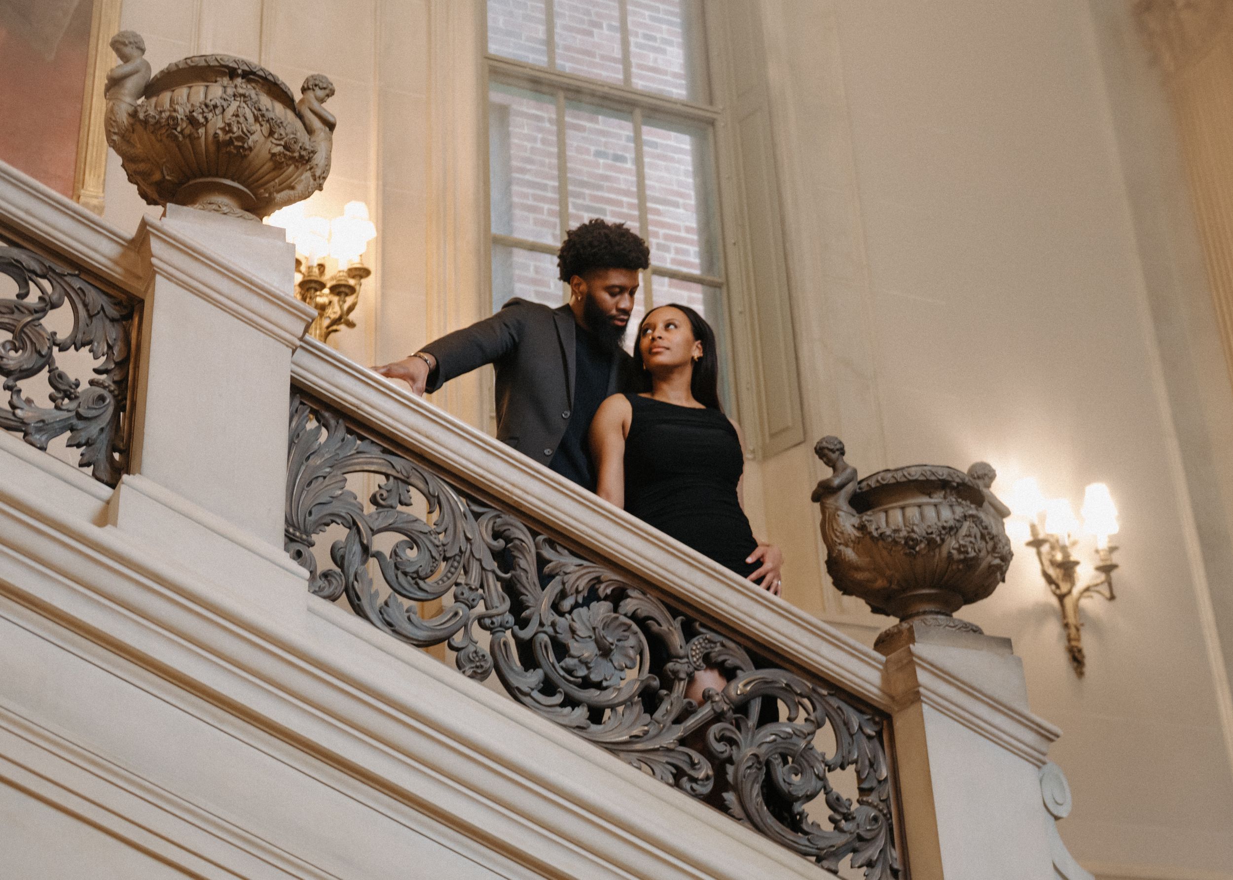 Elegant couple stands on ornate marble staircase with decorative wrought iron railings and classical urns in grand interior.