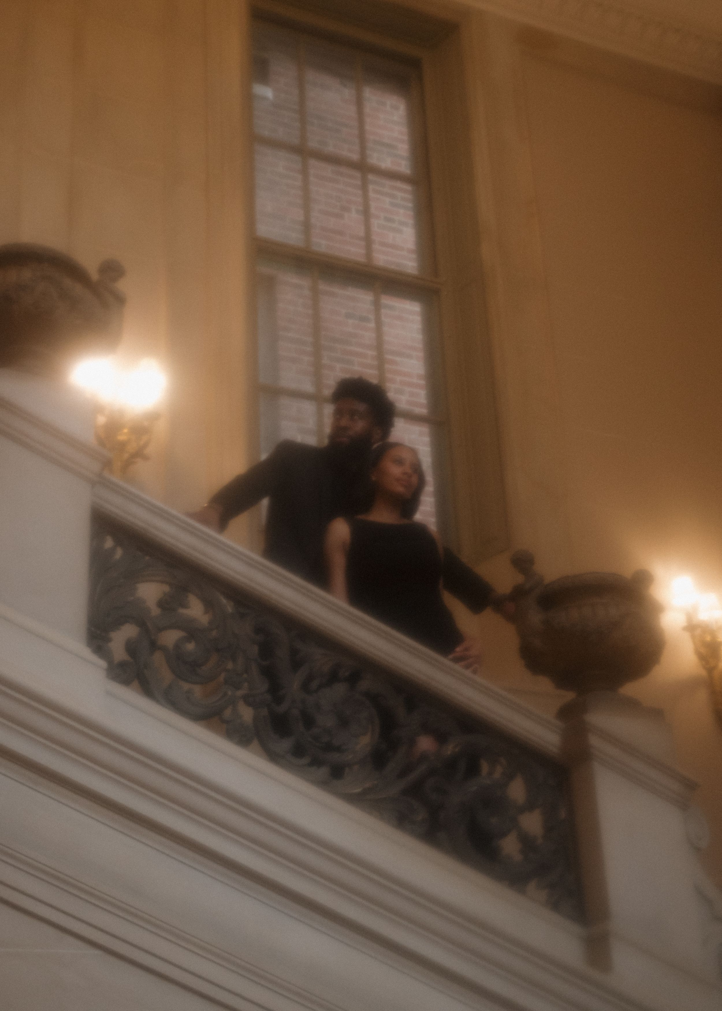 A couple stands together on an elegant staircase with ornate details and warm lighting in an historic interior.