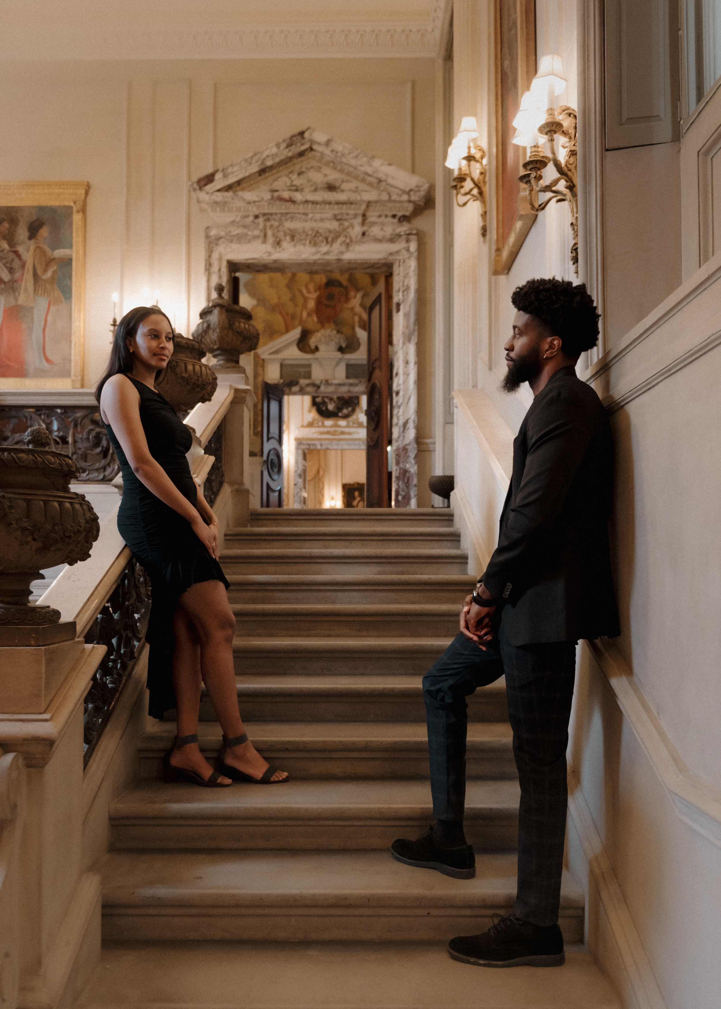 A couple shares a playful moment on an elegant staircase with ornate details and wall sconces in a historic building.