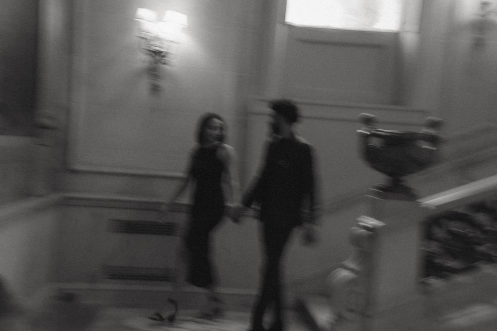 A blurry monochrome photo shows two figures walking down a hallway while holding hands.