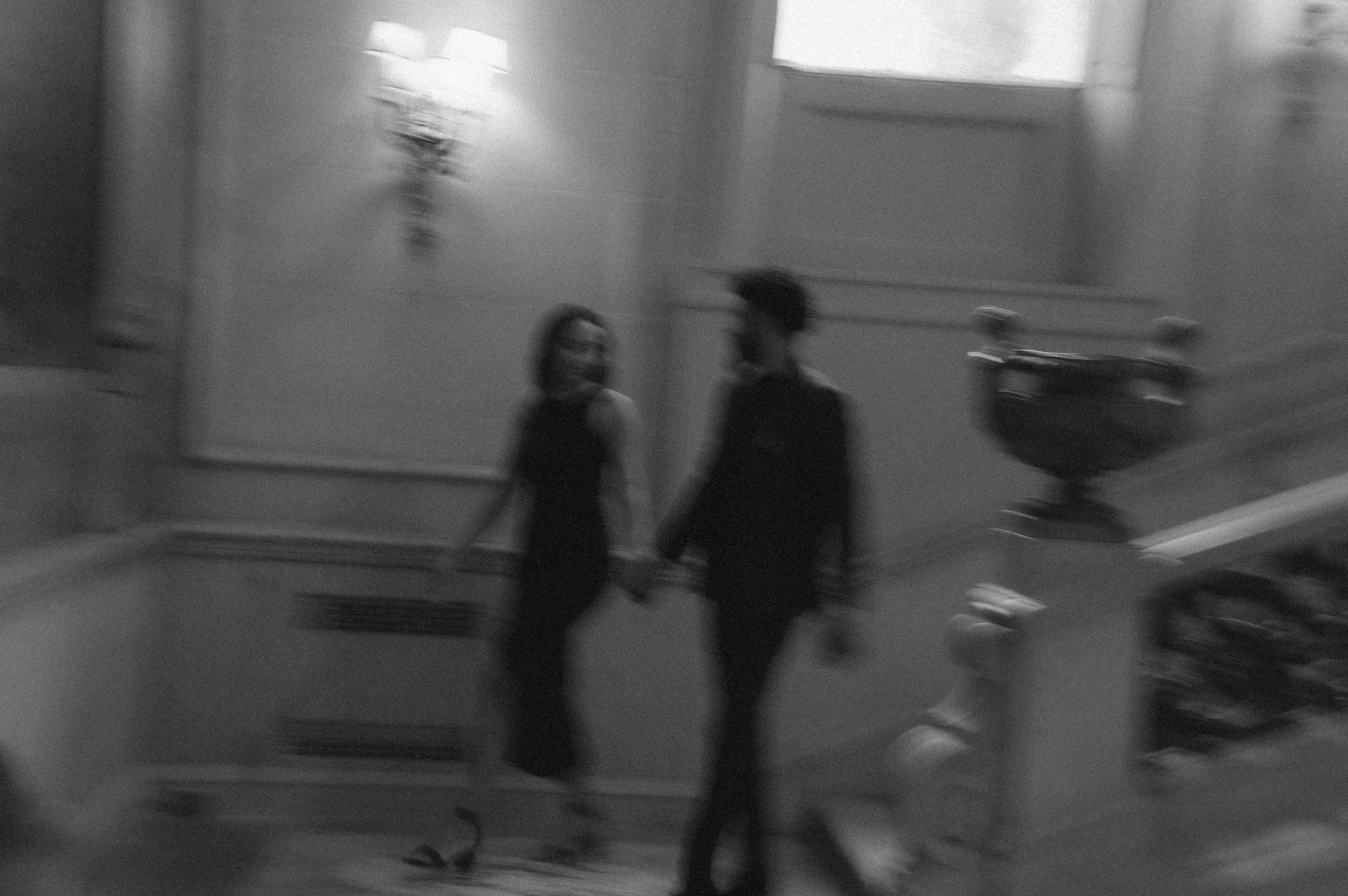 A blurry monochrome photo shows two figures walking down a hallway while holding hands.