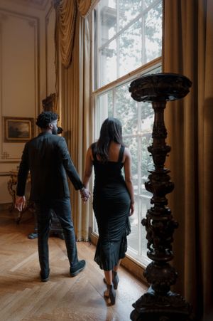 A couple walks hand in hand through an elegant room with tall windows and ornate curtains beside an antique decorative pillar.