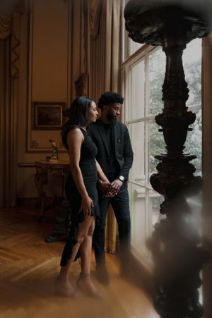A couple in dark attire stands intimately by a grand window in an elegant room with ornate vintage decor.