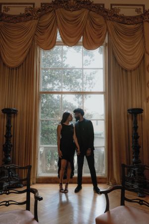 Silhouetted couple stands before an elegant floor-to-ceiling window with ornate golden curtains in a historic room.