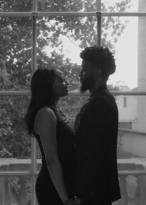 A romantic black and white portrait of a couple standing in front of a large window with trees visible outside.