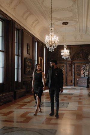 A couple walks hand in hand down an elegant hallway with crystal chandeliers and ornate marble floors in a historic building.