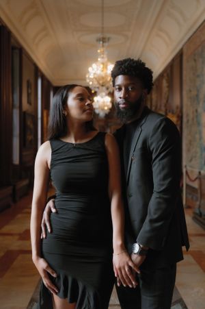 Elegant couple in matching black formal attire pose together in an ornate hallway with crystal chandeliers.